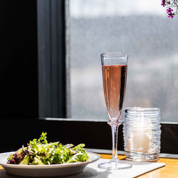 Two clear champagne flutes, one filled with rosé wine, displayed on a table by a window.