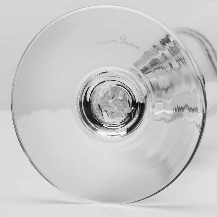 A close-up studio shot of a clear glass wine glass viewed from the base, showing the circular bowl and a faint etched 'Simon Pearce' brand mark on the glass.