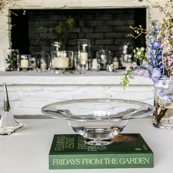 Clear glass bowl with pedestal base displayed on green book, surrounded by flowers and candles.