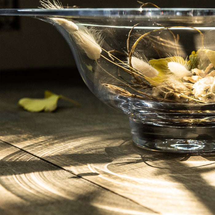 Clear glass bowl filled with dried grasses, wheat, and white botanicals, displayed on wooden surface.