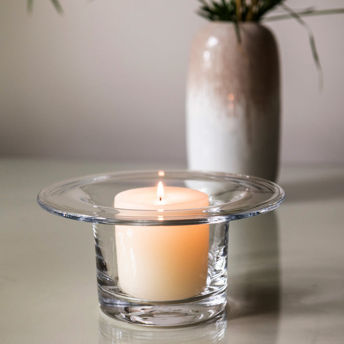Lit cream-colored candle in clear glass hurricane holder with wide rim, displayed with matching vase and greenery in backgrou