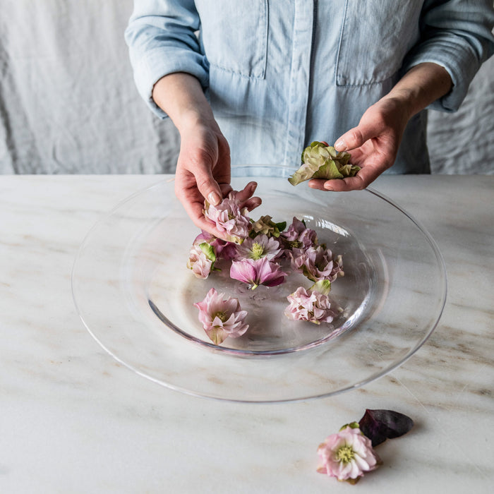 Clear glass platter with person arranging pink and purple edible flowers on marble surface.