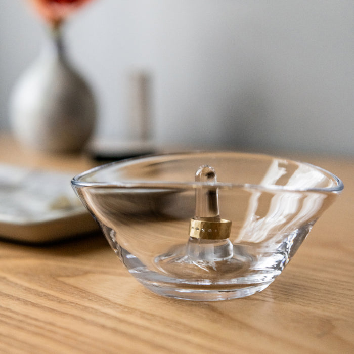 Clear glass bowl with brass and silver central post for holding rings, displayed on wooden surface.
