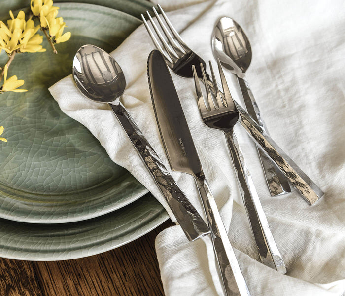 Silver-toned flatware set with fork, knife, and spoons arranged on white napkin next to green plate and yellow flowers.