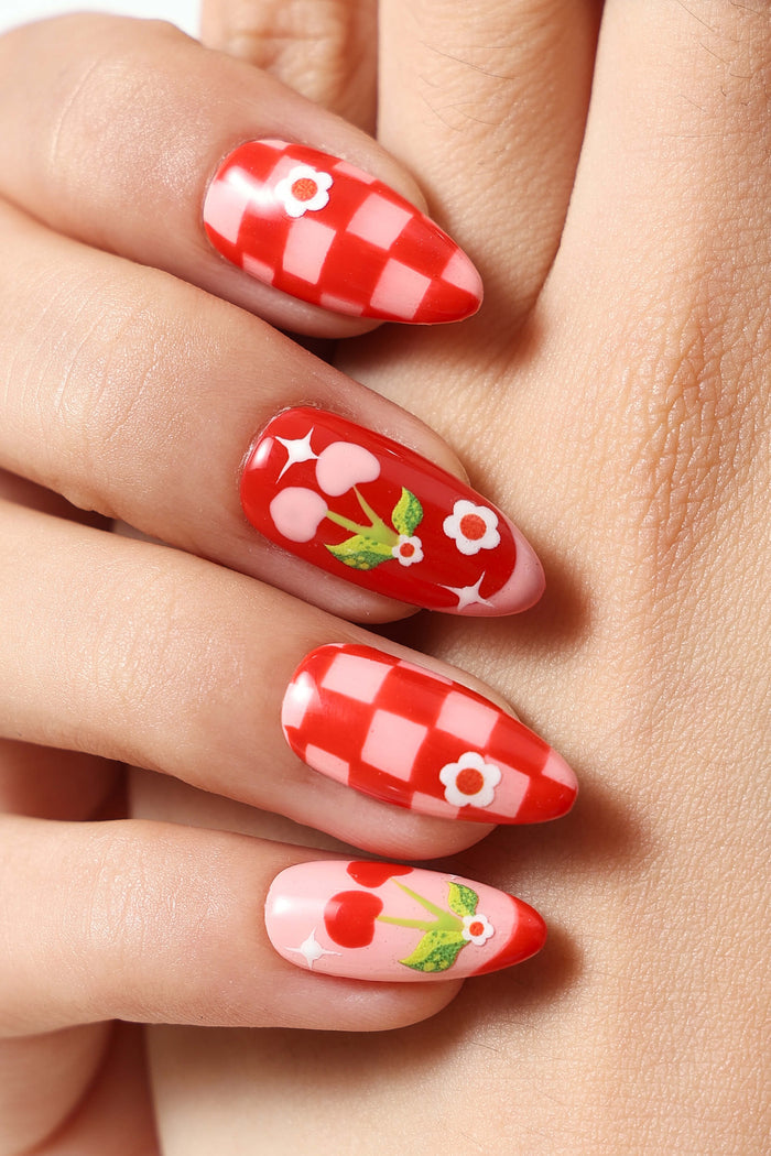 Close-up of a hand with almond-shaped press-on nails featuring a checkered pattern, cherries, and flowers.