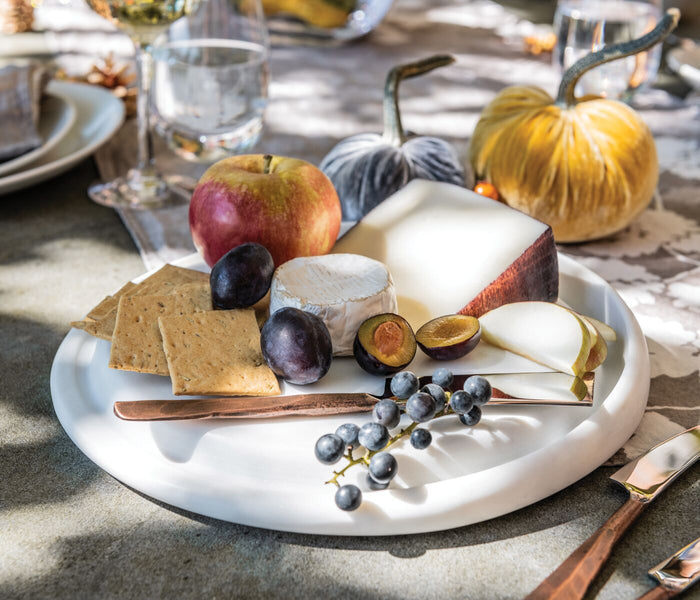 White marble round cheese board with cheese, crackers, grapes, figs, and wooden spreader arranged on top.