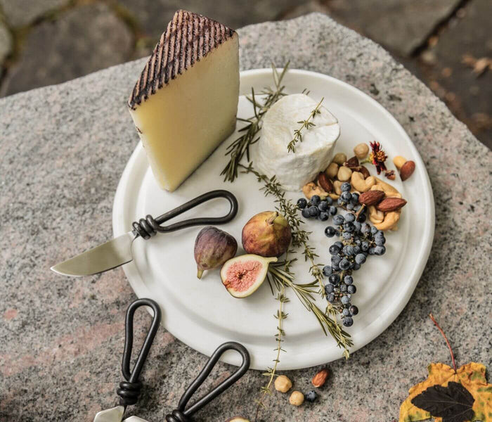 White marble round cheese board with two cheeses, figs, berries, nuts, herbs, and black cheese knives.