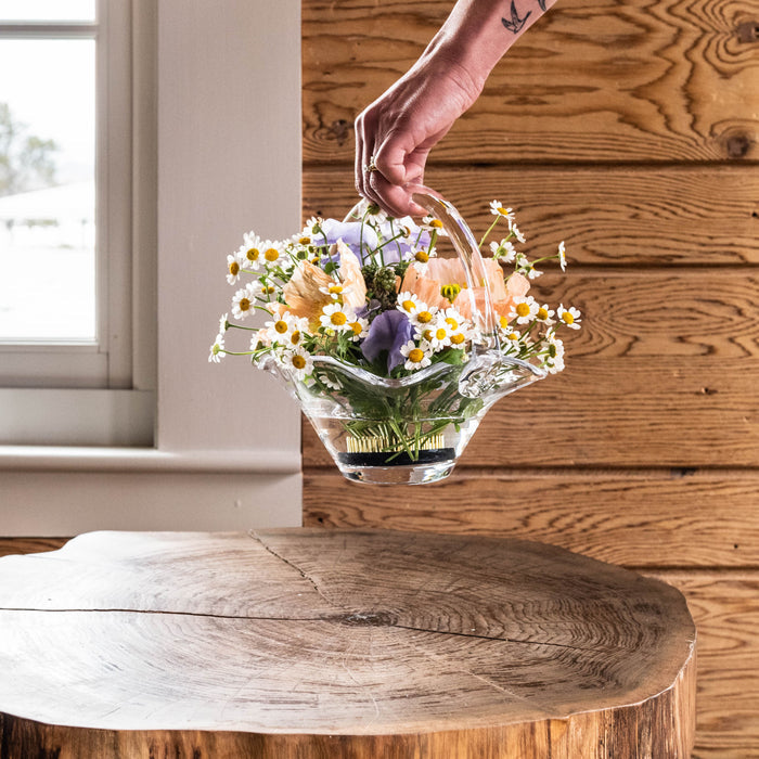 Clear glass basket with handles holding mixed daisies and purple flowers with green stems.