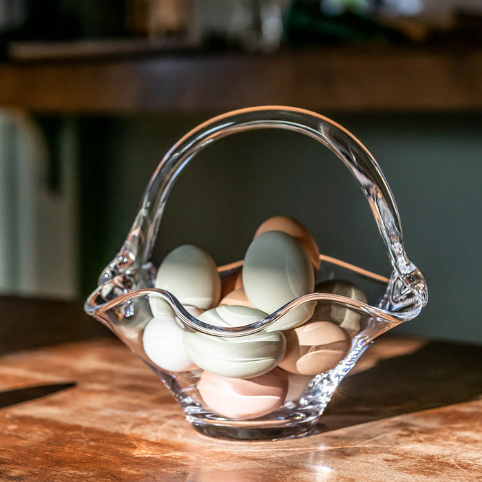 Clear glass basket with wavy rim and handle holding white and brown eggs on wooden surface.