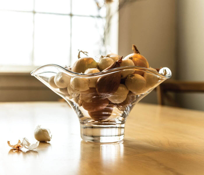 Clear glass bowl with flared ruffled rim containing onions on wooden surface.