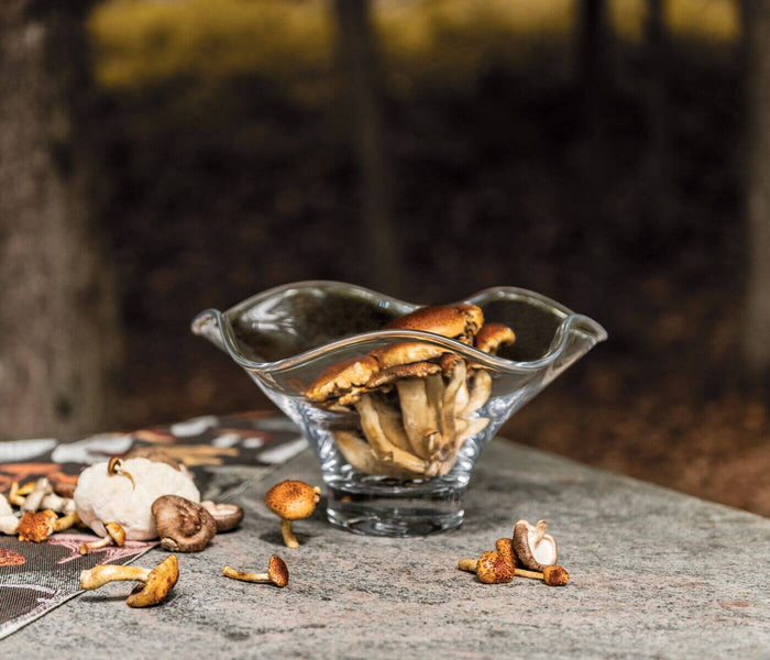 Clear glass bowl with wavy flared rim containing roasted cashews, surrounded by scattered nuts and shells on gray surface.