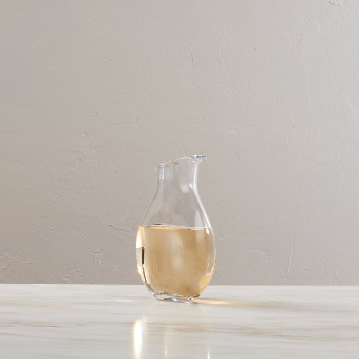 Clear glass carafe with curved body filled with pale liquid, pouring spout at top right, positioned on white surface.