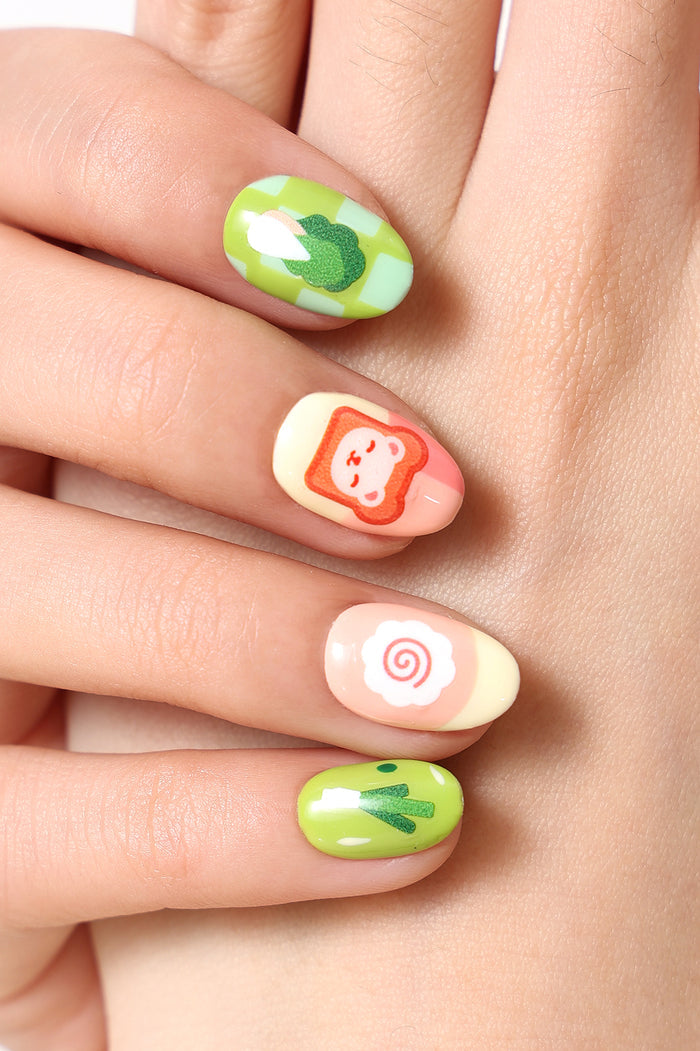 Press-on nails with cute cooking bear, broccoli, and toast designs on a pale pink and yellow gradient.