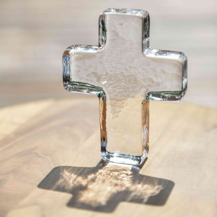Clear glass cross with textured surface and beveled edges on a light wood background.