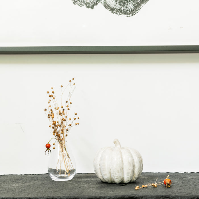 White ridged decorative pumpkin displayed next to dried botanicals in glass vase on shelf.