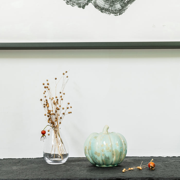 Pale green crystalline pumpkin with textured surface displayed on dark surface next to dried botanicals in glass vase.