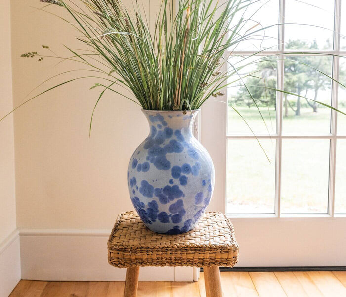 Blue and white ceramic vase with organic pattern, filled with green ornamental grass, displayed on woven stool.