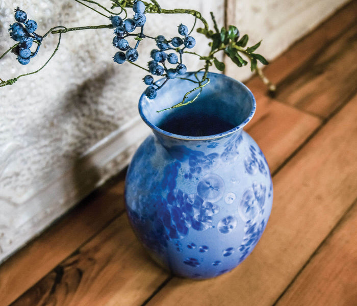 Blue ceramic vase with mottled pattern holding blue berries on green stems, displayed on wooden surface.