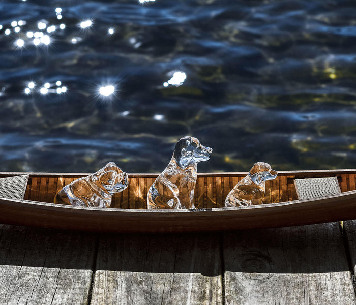 Three clear glass dog figurines arranged on a wooden boat rail with water and reflective light in the background.