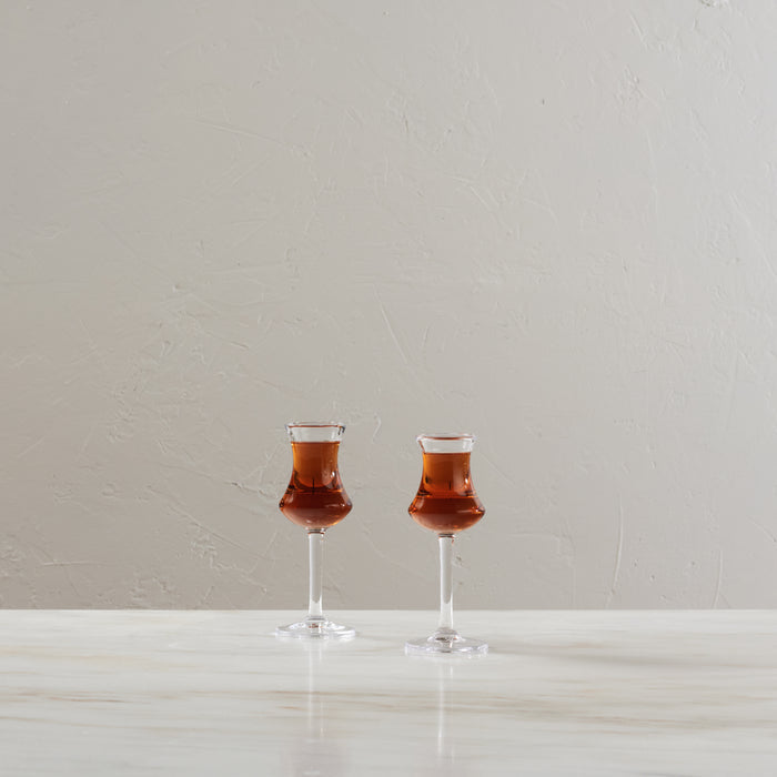 Two clear aperitif glasses filled with reddish-brown liquid, positioned on a white surface against a neutral background.