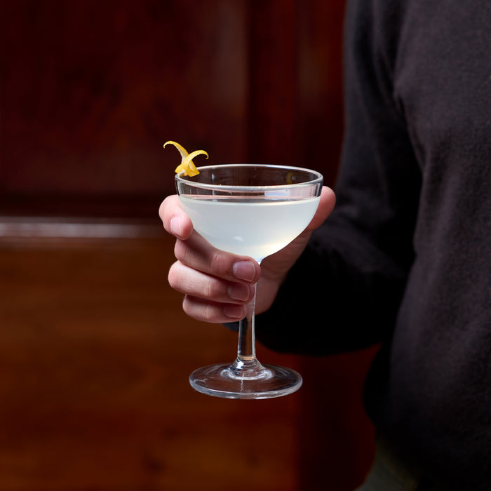 Clear coupe glass with stem filled with pale liquid and yellow citrus twist, held in hand against dark background.