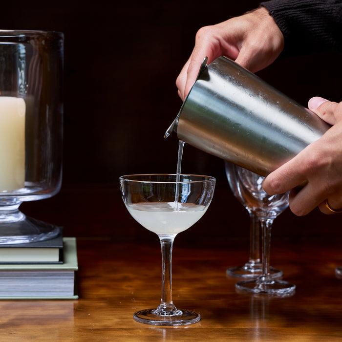 Two coupe glasses with a cocktail being poured from a metal shaker on a wooden surface.