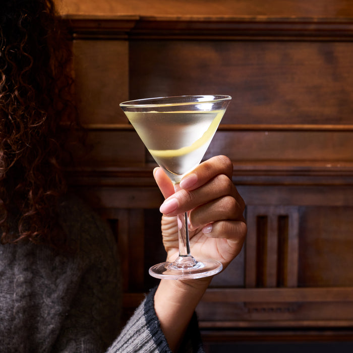 Hand holding a martini glass filled with clear pale yellow liquid against a wood-paneled background.