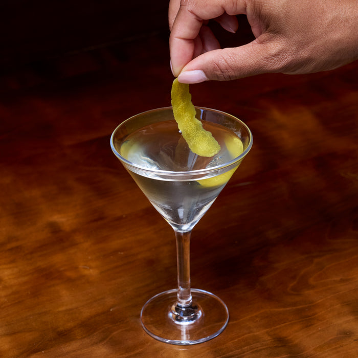 Hand dropping lemon twist into a martini glass filled with clear liquid on a brown surface.