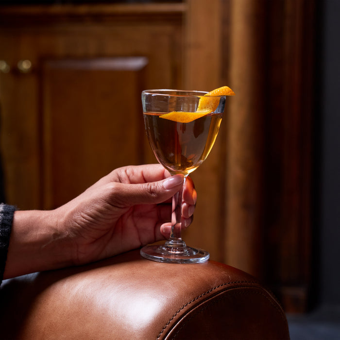 Hand holding a coupe glass with amber liquid and orange peel garnish against a warm interior background.