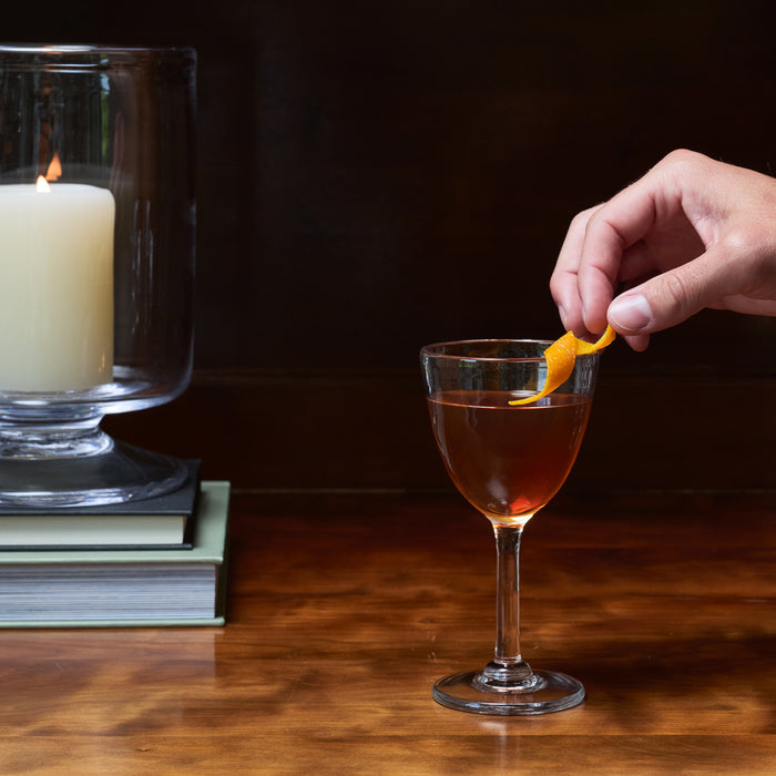 Hand garnishing cocktail glass with orange peel, with second glass containing cream-colored drink in background on wooden sur
