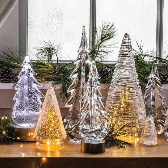 Illuminated decorative tree figurines in frosted and textured finishes with warm white LED lights, arranged with evergreen br
