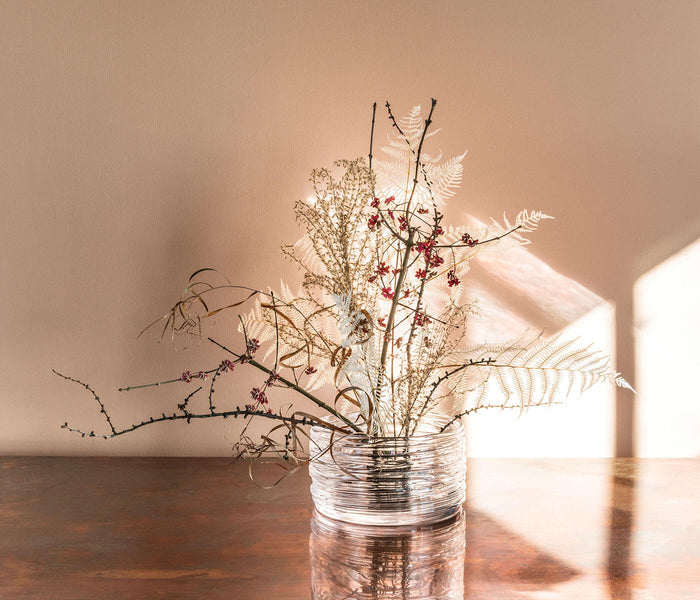 Clear glass bowl with horizontal ridged pattern containing dried flowers, ferns, and branches on wooden surface.