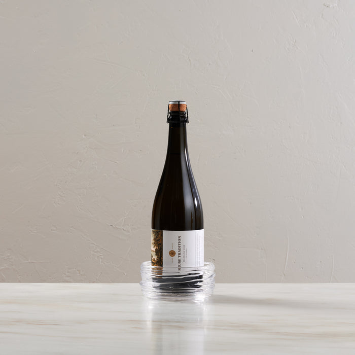 Dark glass bottle with white label sitting in clear glass circular coaster on white surface.