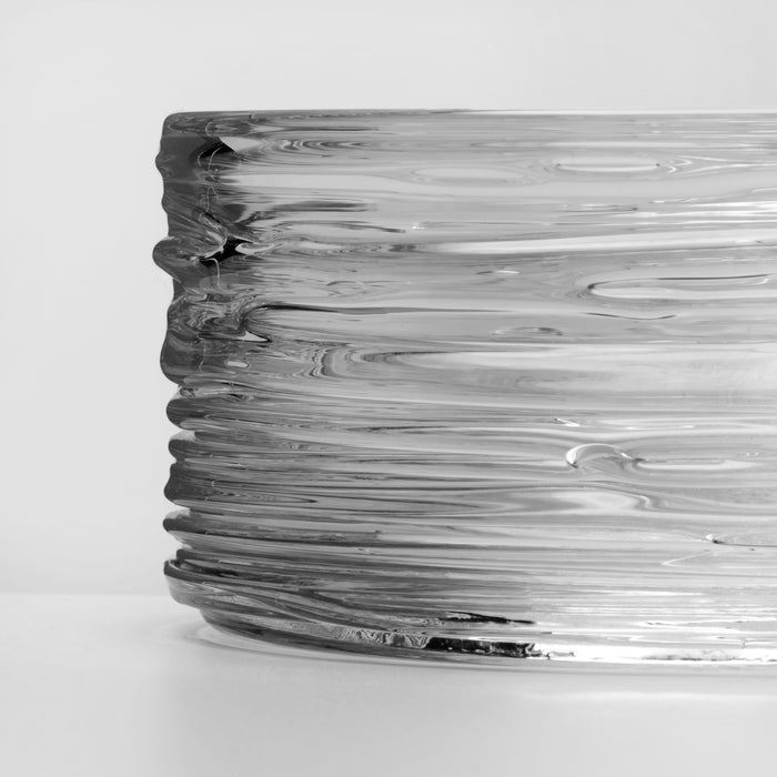 Clear glass coaster with wavy, rippled surface design and dark gray-toned edges.
