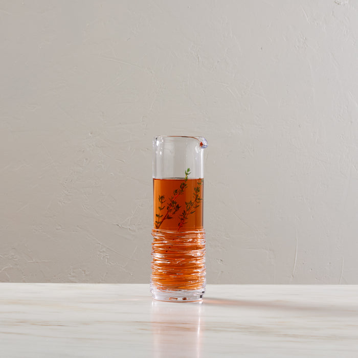 Clear glass carafe filled with amber liquid and herb sprigs, featuring textured lower section and pouring spout.