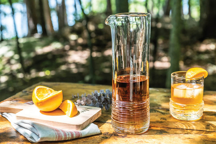 Clear glass carafe with vertical ridged texture containing amber liquid, positioned next to a filled cocktail glass with ice