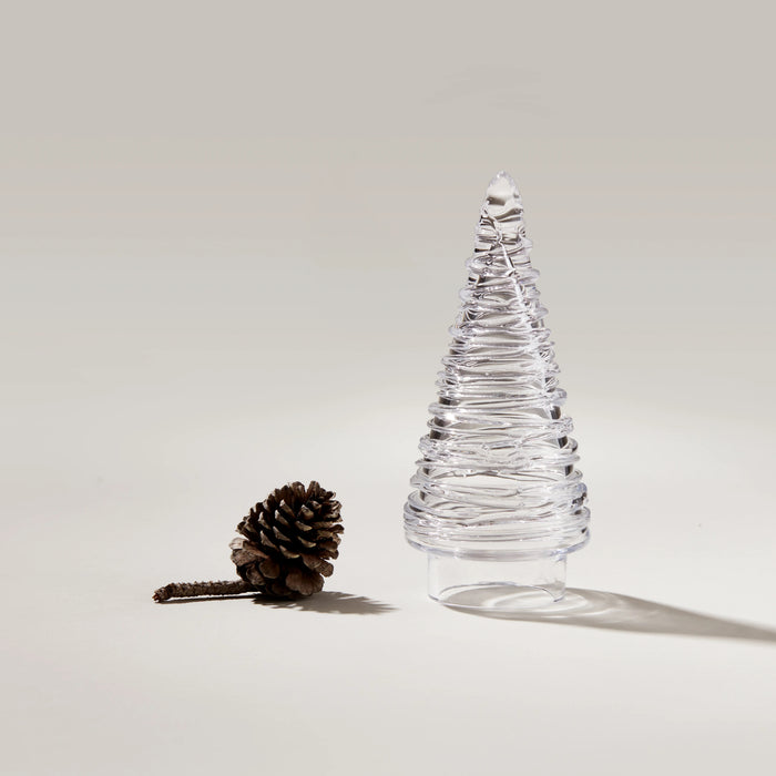 Clear glass tree-shaped container with horizontal ridged bands next to a brown pinecone on neutral background.