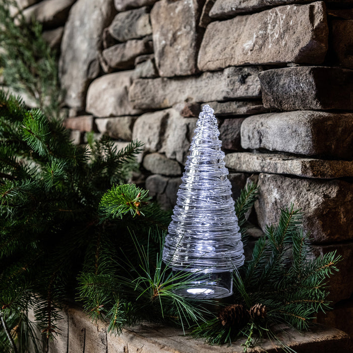 Illuminated wire tree decoration with blue-white lights surrounded by evergreen branches and pinecones against a stone wall.
