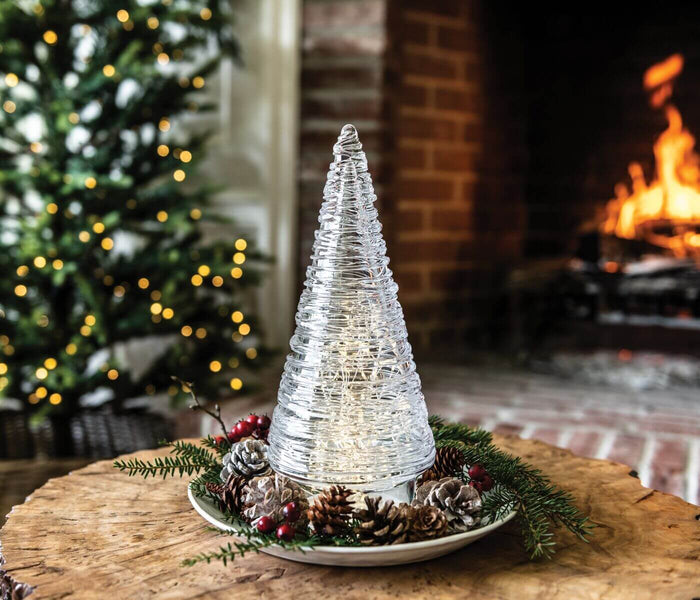 Silver wire-wrapped cone tree with LED lights on ceramic base surrounded by greenery and pinecones.