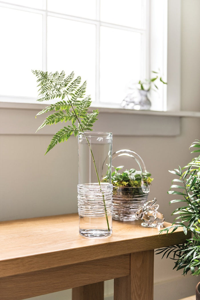 Clear glass cylinder vase with textured lower section, holding green fern fronds on a wooden surface.