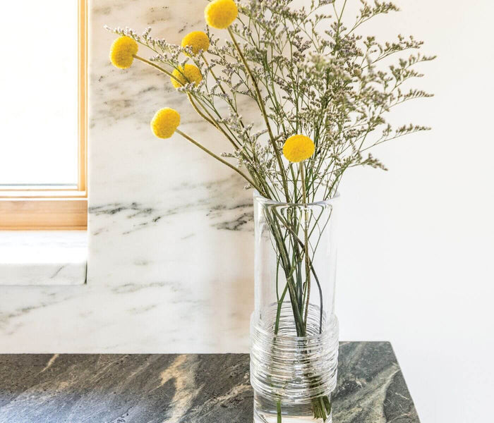 Clear glass vase with horizontal ribbed texture containing yellow ball flowers and green filler stems.
