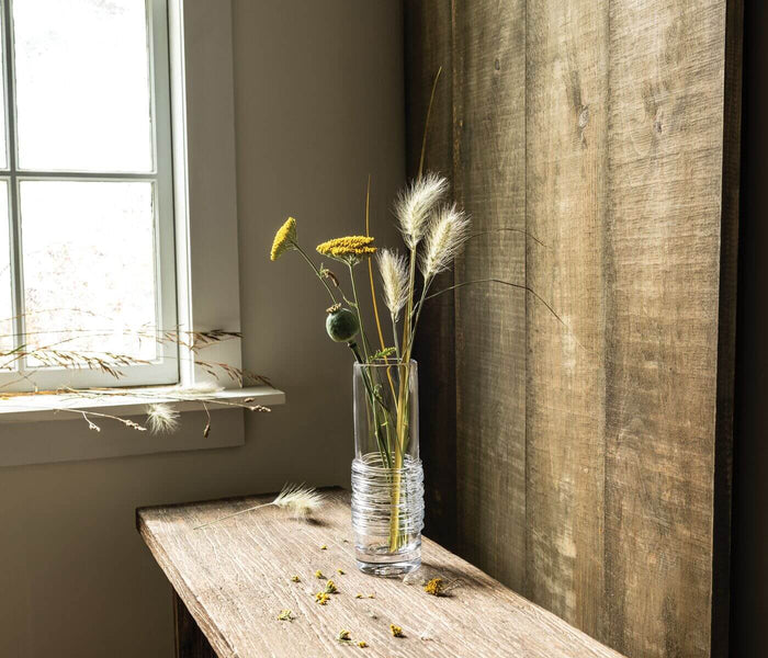 Clear glass vase with dried yellow flowers, white pampas grass, and green seed pods on wooden surface near window.