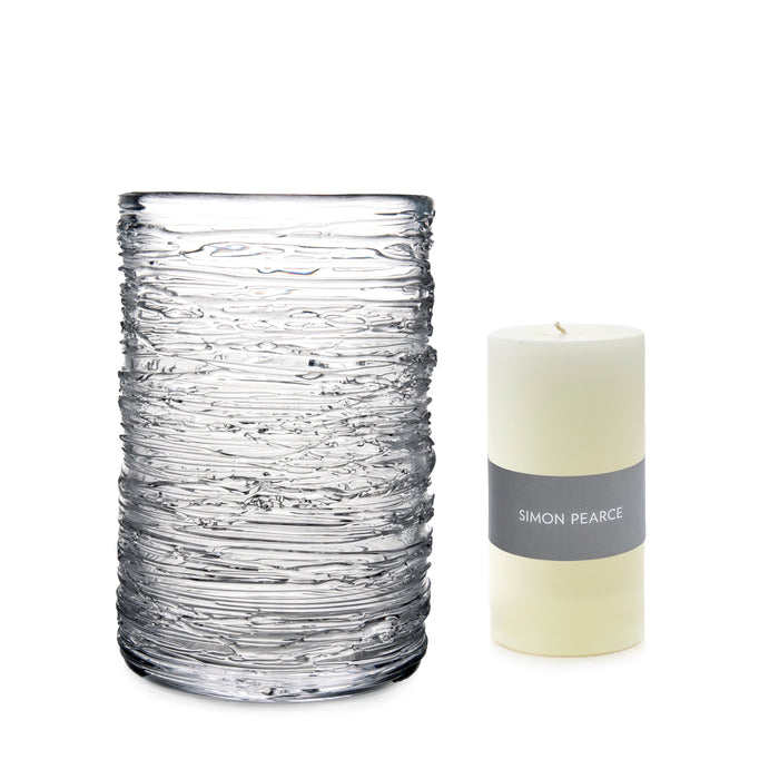 Clear glass hurricane vase with textured surface next to cream pillar candle with gray Simon Pearce band.