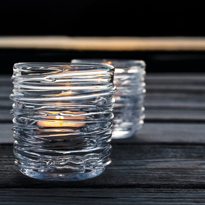 Two clear glass tealight holders with textured wavy design, one lit with yellow flame, on dark wooden surface.