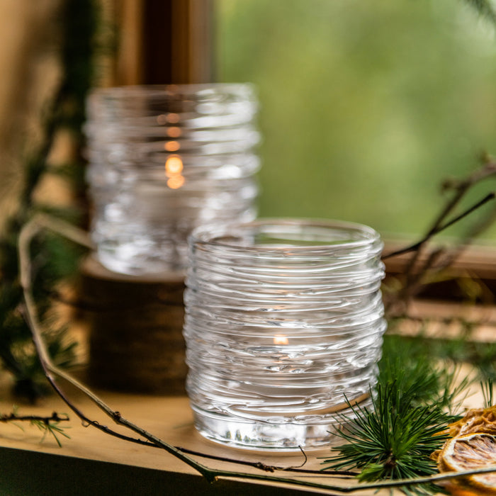 Two clear ribbed glass tealight holders with lit candles, displayed outdoors among evergreen branches and natural decor.