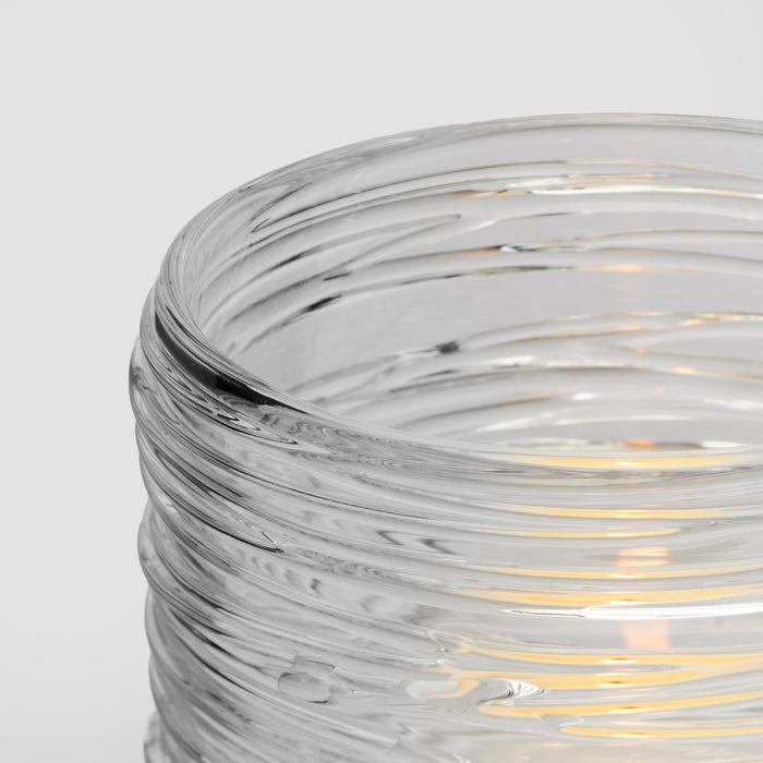Clear glass tealight holder with horizontal ridged texture and lit candle inside.