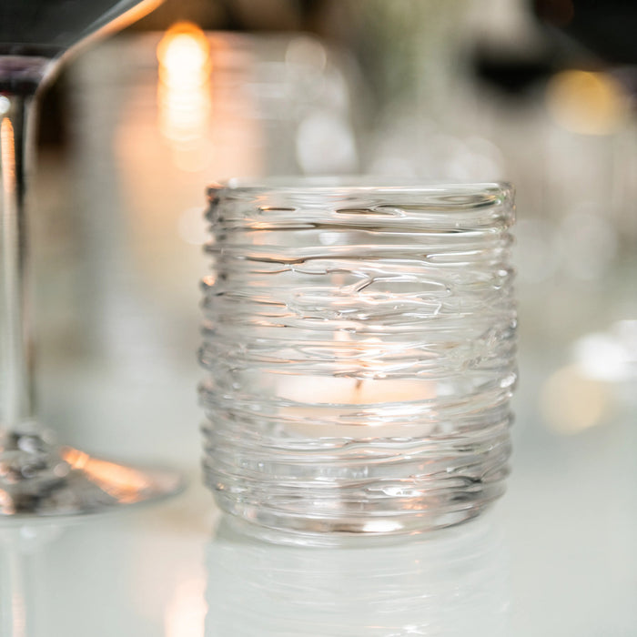 Clear glass tealight holder with horizontal ridged texture and lit candle inside.
