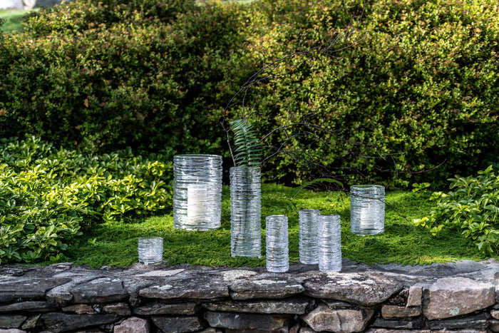 Cylindrical metal vases with horizontal ridged texture in various heights, displayed on stone wall with greenery background.