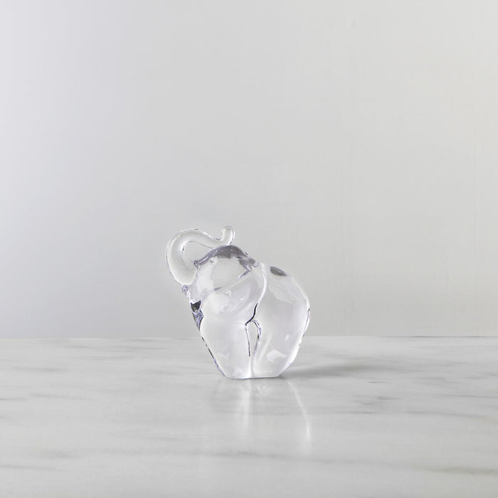 Clear glass elephant figurine with raised trunk on a marble surface against a gray background.