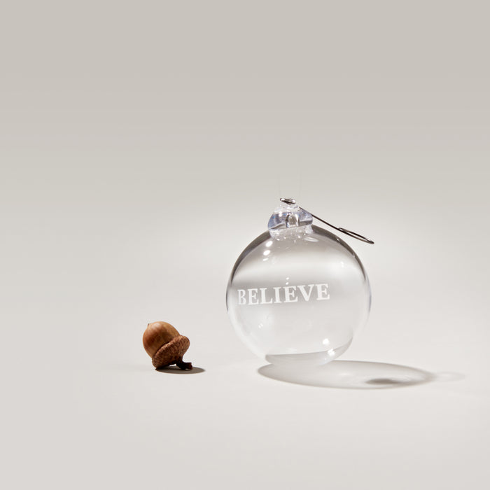 Clear glass round ornament with "BELIEVE" engraved on surface and metal hanging hook.